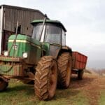 Jak ograniczyć ugniatanie gleby: ogumienie, ciśnienie, balast i ścieżki przejazdowe w praktyce Zielony ciągnik z przyczepą obok starej stodoły w pochmurny dzień